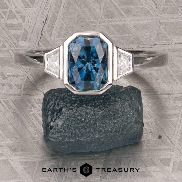 The "Everly" Three-Stone Ring in Platinum 950 with a 1.83-Carat Peacock Blue Montana Sapphire (Heated) The "Everly" Three-Stone Ring in Platinum 950 with a 1.83-Carat Peacock Blue Montana Sapphire (Heated) and two white side stones, displayed on a rough blue stone with the Earth’s Treasury logo below.