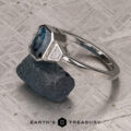 The "Everly" Three-Stone Ring in Platinum 950 with a 1.83-Carat Peacock Blue Montana Sapphire (Heated) flanked by two side stones is showcased atop a rugged dark stone, with a textured metal backdrop and the Earth’s Treasury logo displayed at the bottom.