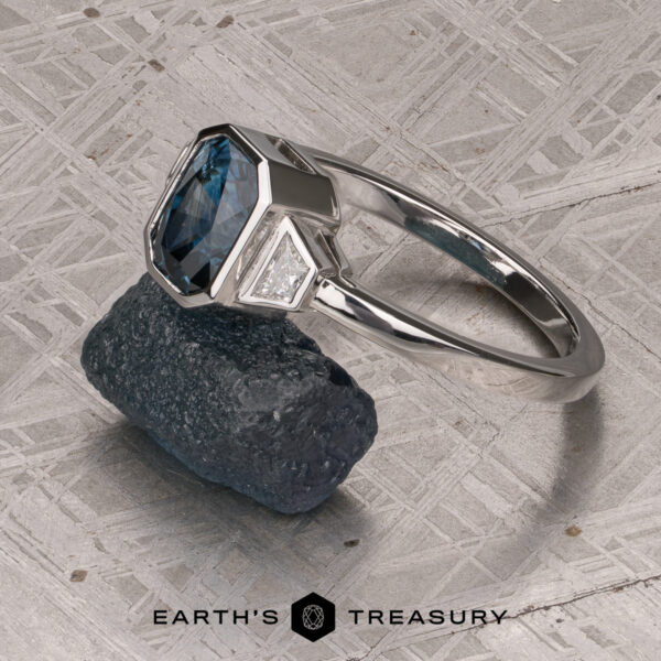 The "Everly" Three-Stone Ring in Platinum 950 with a 1.83-Carat Peacock Blue Montana Sapphire (Heated) The "Everly" Three-Stone Ring in Platinum 950 with a 1.83-Carat Peacock Blue Montana Sapphire (Heated) flanked by two side stones is showcased atop a rugged dark stone, with a textured metal backdrop and the Earth’s Treasury logo displayed at the bottom.