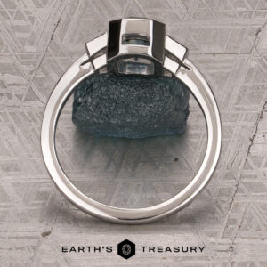 The "Everly" Three-Stone Ring in Platinum 950 with a 1.83-Carat Peacock Blue Montana Sapphire (Heated)—is displayed upright on a textured surface, with “EARTH’S TREASURY” and a geometric logo at the bottom.