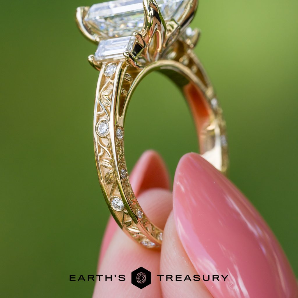 A hand with pink polished nails holds the Custom Ring – Recessed Details Example, featuring exquisite engraving and embedded diamonds that add distinctive texture to its intricate design. The “EARTH’S TREASURY” logo is displayed at the bottom.