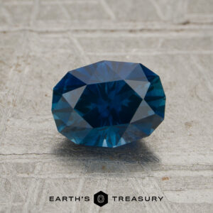 A faceted oval-cut 1.16-carat deep blue Montana sapphire (heated) is placed on a textured gray surface. The text “EARTH’S TREASURY” appears at the bottom of the image.