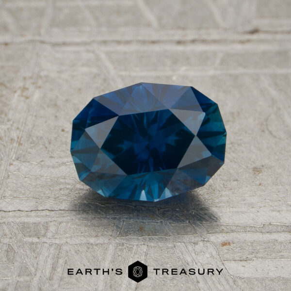 A faceted oval-cut 1.16-carat deep blue Montana sapphire (heated) is placed on a textured gray surface. The text “EARTH’S TREASURY” appears at the bottom of the image.