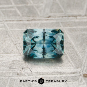A 0.89-carat heated Montana sapphire with a rectangular shape and blue-green hue, featuring geometric facets, rests on a textured metallic surface. The Earth’s Treasury logo appears at the bottom of the image.