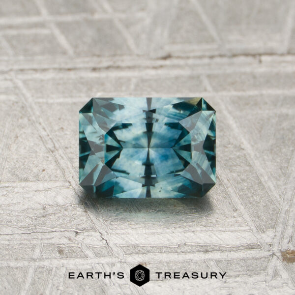 0.89-Carat Teal Montana Sapphire (Heated) A 0.89-carat heated Montana sapphire with a rectangular shape and blue-green hue, featuring geometric facets, rests on a textured metallic surface. The Earth’s Treasury logo appears at the bottom of the image.