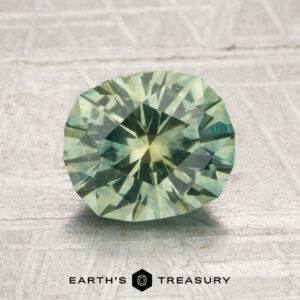 A 1.92-carat heated Montana sapphire, expertly faceted in an oval cut and displaying a pale green hue, is showcased on a textured gray surface with the “EARTH’S TREASURY” logo and text beneath it.