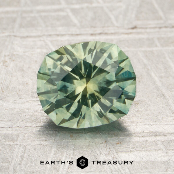 1.92-Carat Mint Green Montana Sapphire (Heated) A 1.92-carat heated Montana sapphire, expertly faceted in an oval cut and displaying a pale green hue, is showcased on a textured gray surface with the “EARTH’S TREASURY” logo and text beneath it.