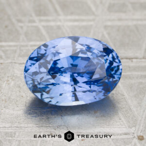 A 4.82-carat cornflower blue to violet color-change Ceylon sapphire rests on a textured light gray surface, with the text “EARTH’S TREASURY” and a geometric logo displayed below the gem.