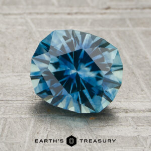 A 1.98-carat teal blue Montana sapphire (heated) with a round brilliant cut is displayed on a textured gray surface, with the “EARTH’S TREASURY” text and logo beneath it.
