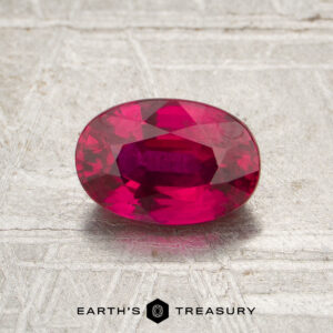 A 1.10-carat heated Thai ruby, cut in an oval shape, rests on a textured gray surface. The text “EARTH’S TREASURY” appears at the bottom of the image.
