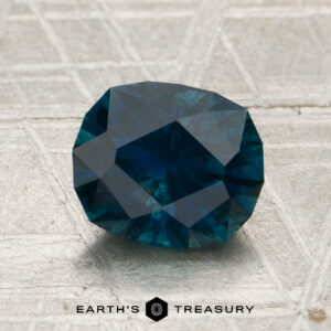 A 2.02-carat deep peacock blue Montana sapphire (heated), intricately faceted, rests on a textured, light-colored surface. The Earth’s Treasury logo is displayed at the bottom of the image.