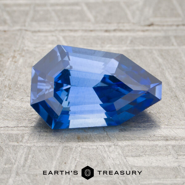 1.84-Carat Cornflower Blue Ceylon Sapphire (Heated) A 1.84-carat cornflower blue Ceylon sapphire (heated) rests on a light textured surface, showcasing sharp geometric angles and clear reflections. The text “EARTH’S TREASURY” appears at the bottom.