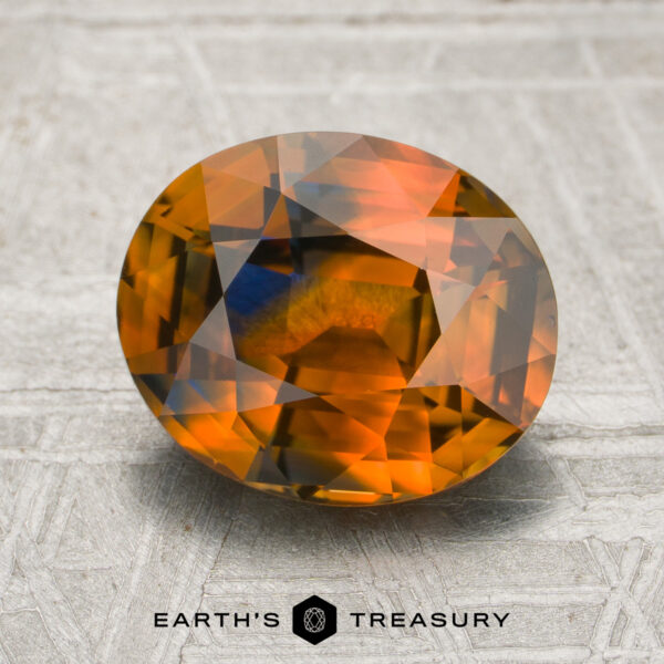 3.04-Carat Orange-Blue Particolored Ceylon Sapphire (Heated) A 3.04-carat orange-blue particolored sapphire (heated) in an oval cut, showcasing vibrant orange, yellow, and blue hues on a textured gray surface; the “Earth’s Treasury” logo and text appear at the bottom.