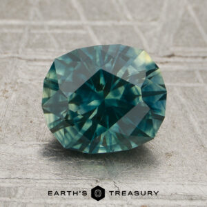 A 1.19-carat deep teal green-yellow particolored Montana sapphire, cut in an oval faceted shape, is displayed on a textured gray surface with the text “EARTH’S TREASURY” and a hexagonal logo at the bottom.