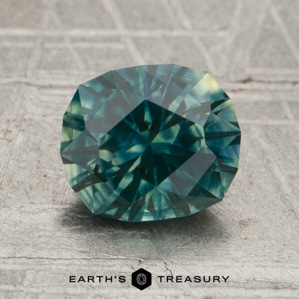 1.91-Carat Deep Teal Green-Yellow Particolored Montana Sapphire (Heated) A 1.19-carat deep teal green-yellow particolored Montana sapphire, cut in an oval faceted shape, is displayed on a textured gray surface with the text “EARTH’S TREASURY” and a hexagonal logo at the bottom.