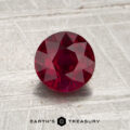 A round, faceted 1.14-carat Mozambique ruby (heated) rests at the center of a textured gray surface, with the words “EARTH’S TREASURY” displayed at the bottom of the image.