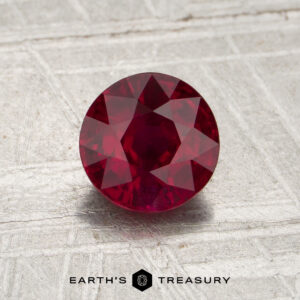 A round, faceted 1.14-carat Mozambique ruby (heated) rests at the center of a textured gray surface, with the words “EARTH’S TREASURY” displayed at the bottom of the image.