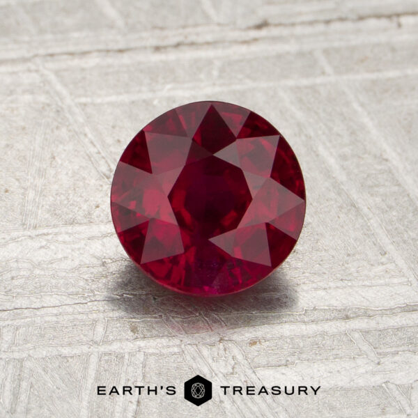 1.14-Carat Mozambique Ruby (Heated) A round, faceted 1.14-carat Mozambique ruby (heated) rests at the center of a textured gray surface, with the words “EARTH’S TREASURY” displayed at the bottom of the image.