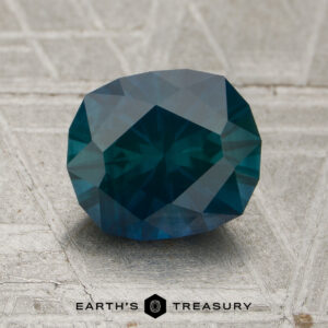 A 1.87-carat deep blue-green Montana sapphire (heated) is displayed on a textured light-colored surface with the text “EARTH’S TREASURY” below.
