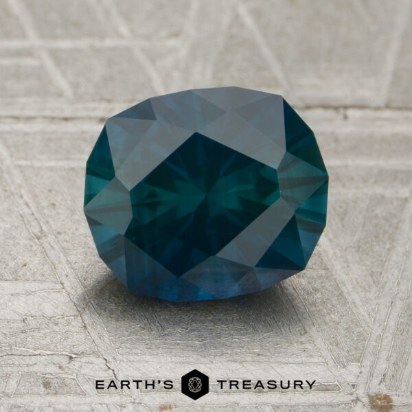 1.87-Carat Deep Blue-Green Montana Sapphire (Heated) A 1.87-carat deep blue-green Montana sapphire (heated) is displayed on a textured light-colored surface with the text “EARTH’S TREASURY” below.