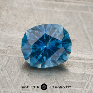 A 1.67-carat heated Montana sapphire rests on a textured beige surface, with the words “EARTH’S TREASURY” and a hexagon logo displayed below.