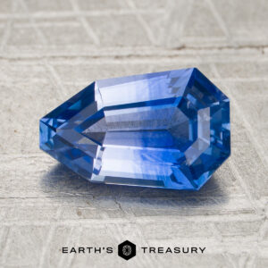 A 2.00-carat rich blue-colourless particolored Ceylon sapphire with geometric facets rests on a textured surface. The logo and text “Earth’s Treasury” appear at the bottom.