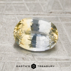 2.13-carat colorless-yellow particolored Ceylon sapphire (heated), oval-cut and showcased against a textured gray background with Earth’s Treasury branding at the bottom, featuring striking yellow and blue hues.
