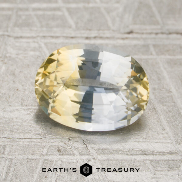 2.13-Carat Colorless-Yellow Particolored Ceylon Sapphire (Heated 2.13-carat colorless-yellow particolored Ceylon sapphire (heated), oval-cut and showcased against a textured gray background with Earth’s Treasury branding at the bottom, featuring striking yellow and blue hues.