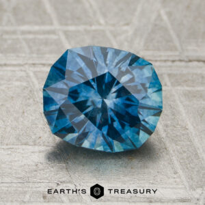 A 1.80-carat medium blue Montana sapphire (heated) rests on a textured gray surface, with the Earth’s Treasury logo and text visible at the bottom of the image.
