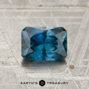 A 1.32-carat heated Montana sapphire, rectangular and faceted in shape with a deep blue hue, rests on a textured gray surface. The logo “EARTH’S TREASURY” appears at the bottom of the image.