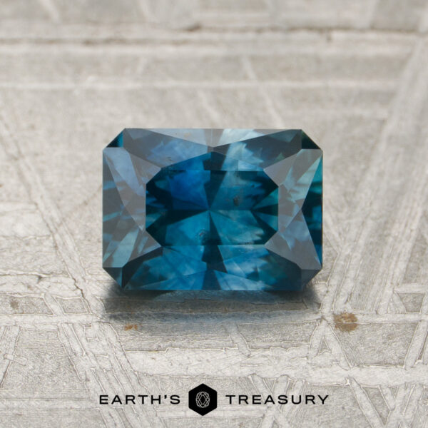 1.32-Carat Peacock Blue Montana Sapphire (Heated) A 1.32-carat heated Montana sapphire, rectangular and faceted in shape with a deep blue hue, rests on a textured gray surface. The logo “EARTH’S TREASURY” appears at the bottom of the image.