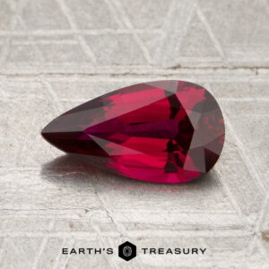 A faceted, pear-shaped 1.31-carat Mozambique Ruby (heated) in deep red rests on a textured, metallic surface with the “Earth’s Treasury” logo at the bottom.