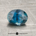 A 1.57-carat teal Madagascar sapphire, cut in an oval shape with reflective facets, rests on a textured gray surface. The text “Earth’s Treasury” appears at the bottom of the image.