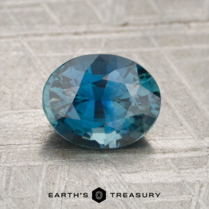 A 1.57-carat teal Madagascar sapphire, cut in an oval shape with reflective facets, rests on a textured gray surface. The text “Earth’s Treasury” appears at the bottom of the image.