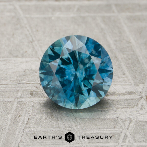 1.25-carat teal blue Montana sapphire (heated) with multiple facets displayed on a textured surface, featuring the words “EARTH’S TREASURY” and a hexagonal logo below.