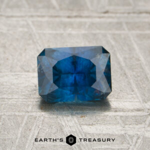 A 1.14-carat Montana sapphire (heated), cut in a rectangular shape with multiple facets and displaying a rich blue hue, rests on a textured, light-colored surface. The text “EARTH’S TREASURY” is shown at the bottom.