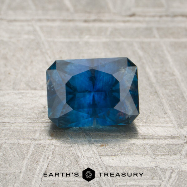 1.14-Carat Deep Blue Montana Sapphire (Heated) A 1.14-carat Montana sapphire (heated), cut in a rectangular shape with multiple facets and displaying a rich blue hue, rests on a textured, light-colored surface. The text “EARTH’S TREASURY” is shown at the bottom.