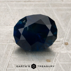A 2.54-carat midnight blue Montana sapphire (heated), cut in a faceted oval shape, rests on a textured light gray surface, with the “Earth’s Treasury” name and hexagon logo visible at the bottom of the image.