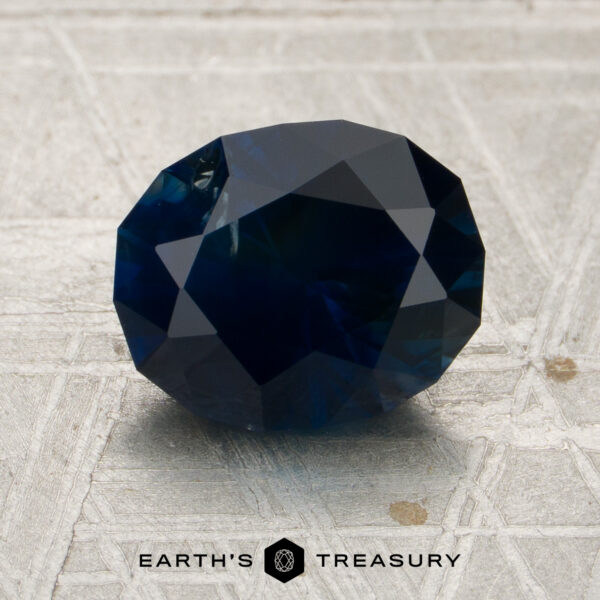 2.54-Carat Near-Black Midnight Blue Montana Sapphire (Heated) A 2.54-carat midnight blue Montana sapphire (heated), cut in a faceted oval shape, rests on a textured light gray surface, with the “Earth’s Treasury” name and hexagon logo visible at the bottom of the image.
