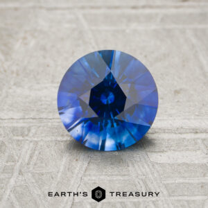 A 1.13-carat royal blue Ceylon sapphire (heated), cut in a round brilliant style, sits on a textured surface, with the text “EARTH’S TREASURY” and a hexagonal logo below.