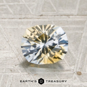 A faceted oval 1.53-carat icy blue and pale yellow particolored Montana sapphire rests on a textured gray surface, with the “Earth’s Treasury” logo below.