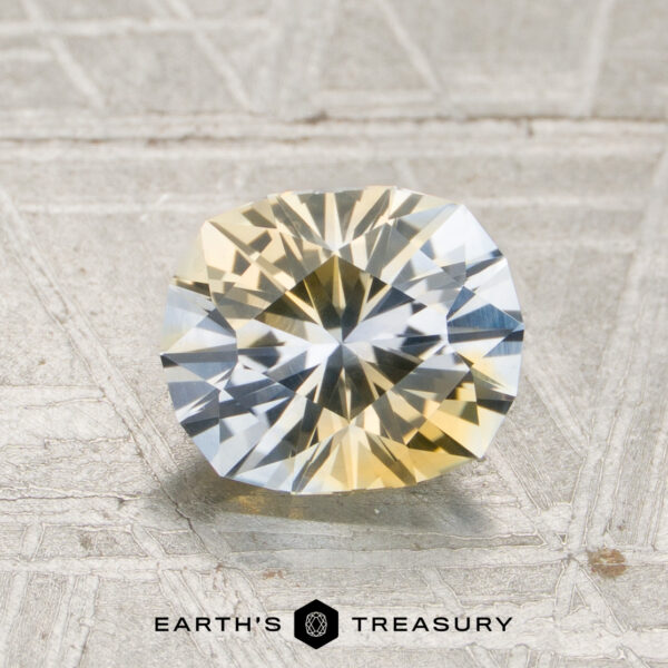 1.53-Carat Icy Blue-Pale Yellow Particolored Montana Sapphire (Heated) A faceted oval 1.53-carat icy blue and pale yellow particolored Montana sapphire rests on a textured gray surface, with the “Earth’s Treasury” logo below.