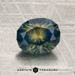 A 2.30-carat deep blue-yellow particolored Montana sapphire, cut in a faceted oval shape, rests on a textured, light-colored surface. “Earth’s Treasury” appears at the bottom of the image.