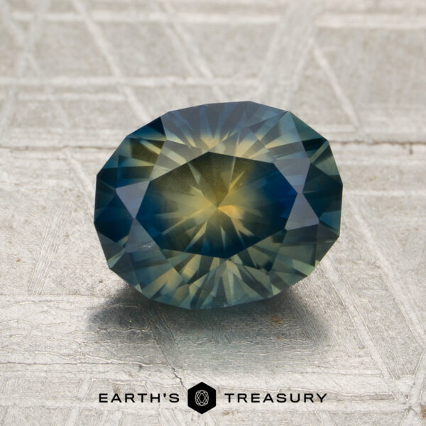 2.30-Carat Deep Blue-Yellow Particolored Montana Sapphire (Heated) A 2.30-carat deep blue-yellow particolored Montana sapphire, cut in a faceted oval shape, rests on a textured, light-colored surface. “Earth’s Treasury” appears at the bottom of the image.