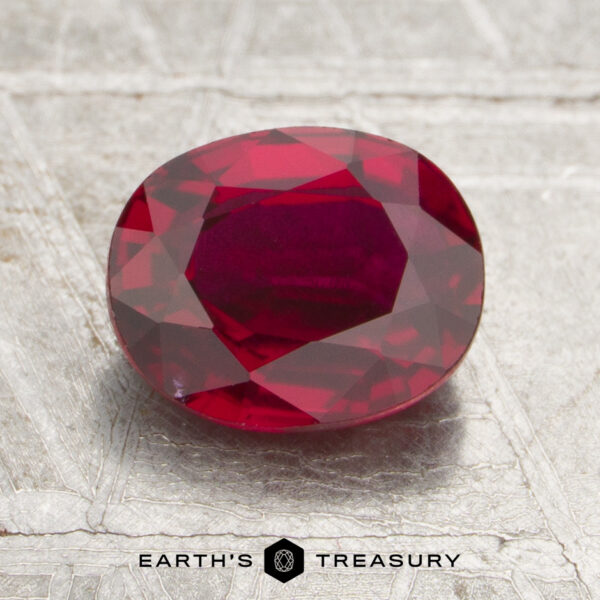 1.29-Carat Mozambique Ruby (Heated) A 1.29-carat Mozambique Ruby (heated), faceted in an oval shape, rests on a textured silver surface, with the Earth’s Treasury logo and text below.