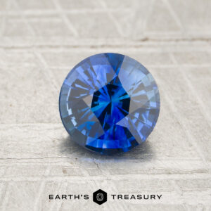 A 1.25-carat royal blue Ceylon sapphire (heated), expertly cut in a round brilliant shape, is displayed on a textured gray surface with the “Earth’s Treasury” logo at the bottom.