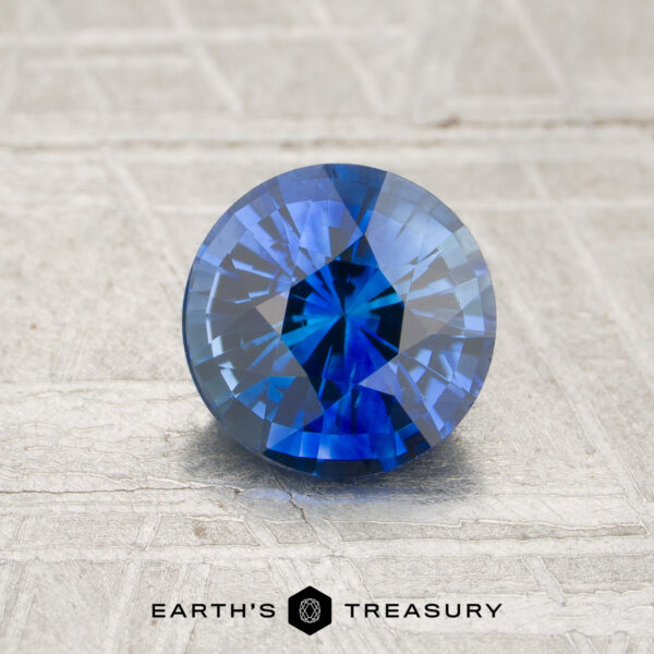 1.25-Carat Royal Blue Ceylon Sapphire (Heated) A 1.25-carat royal blue Ceylon sapphire (heated), expertly cut in a round brilliant shape, is displayed on a textured gray surface with the “Earth’s Treasury” logo at the bottom.