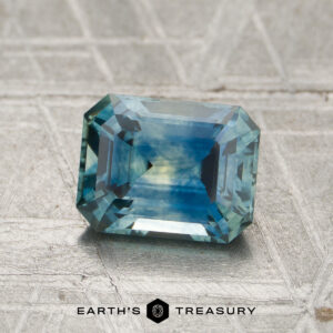 A rectangular step-cut 1.53-carat Montana sapphire with a blue-green hue rests on a textured gray surface, accompanied by the text “EARTH’S TREASURY” and a geometric logo below.