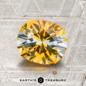 A 2.00-carat heated Montana sapphire, featuring round facets and yellow-blue hues, is displayed on a textured gray surface with the text “Earth’s Treasury” below.
