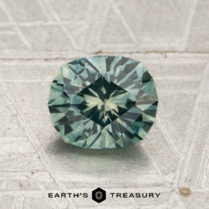 A 1.44-carat Spruce Green Montana Sapphire (heated), featuring angular faceting and a greenish-blue hue, is displayed on a metallic, textured surface above the Earth’s Treasury logo.